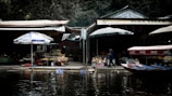 A riverside marketplace featuring a variety of stalls with items displayed for sale. Large umbrellas provide shade over the merchandise. Wooden boats with Coca-Cola branding are docked by the water, and a person is walking along the area. The atmosphere appears vibrant with various colors and textures of goods on display.