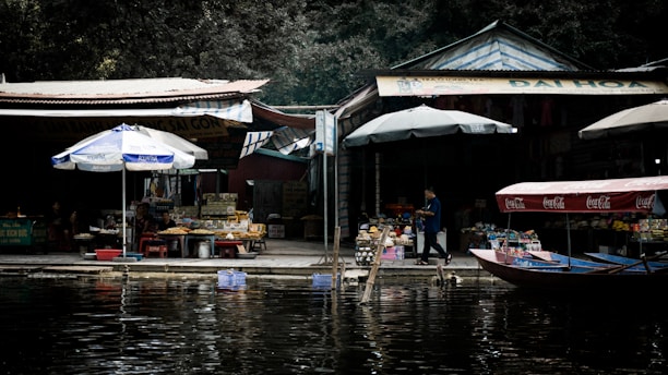 A riverside marketplace featuring a variety of stalls with items displayed for sale. Large umbrellas provide shade over the merchandise. Wooden boats with Coca-Cola branding are docked by the water, and a person is walking along the area. The atmosphere appears vibrant with various colors and textures of goods on display.