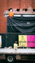 A construction worker wearing an orange outfit and white helmet stands on a platform against a red brick wall. The wall has some holes and a black tarp partially covering it. Below, there is a person in a yellow safety vest looking at colorful posters on a black temporary wall. The posters are bright with bold text and graphics, featuring the words 'RITON' and details about an event. In front of the wall, a truck with construction materials is parked.