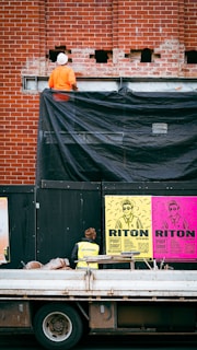 A construction worker wearing an orange outfit and white helmet stands on a platform against a red brick wall. The wall has some holes and a black tarp partially covering it. Below, there is a person in a yellow safety vest looking at colorful posters on a black temporary wall. The posters are bright with bold text and graphics, featuring the words 'RITON' and details about an event. In front of the wall, a truck with construction materials is parked.