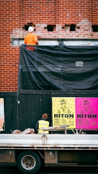 A construction worker wearing an orange outfit and white helmet stands on a platform against a red brick wall. The wall has some holes and a black tarp partially covering it. Below, there is a person in a yellow safety vest looking at colorful posters on a black temporary wall. The posters are bright with bold text and graphics, featuring the words 'RITON' and details about an event. In front of the wall, a truck with construction materials is parked.