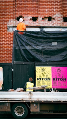 A construction worker wearing an orange outfit and white helmet stands on a platform against a red brick wall. The wall has some holes and a black tarp partially covering it. Below, there is a person in a yellow safety vest looking at colorful posters on a black temporary wall. The posters are bright with bold text and graphics, featuring the words 'RITON' and details about an event. In front of the wall, a truck with construction materials is parked.