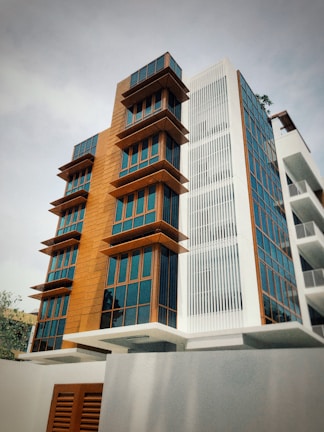 A modern multi-story building with a combination of wooden panels and large glass windows. The structure features angular balconies and a mix of textures with vertical white slats on one segment.