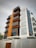 A modern multi-story building with a combination of wooden panels and large glass windows. The structure features angular balconies and a mix of textures with vertical white slats on one segment.