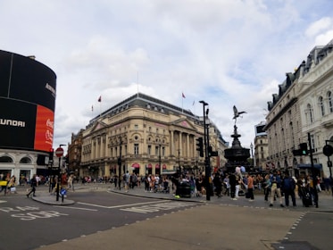 A busy urban intersection with a large digital advertisement screen on the left, displaying multiple brand logos. An ornate, historical building stands in the center, with an iconic statue on a pedestal. Numerous pedestrians are visible crossing the streets and gathering around the statue. The architectural design of the surrounding buildings is classical, and the sky is partly cloudy.