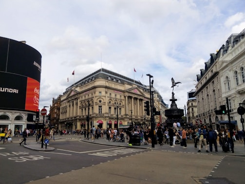 A busy urban intersection with a large digital advertisement screen on the left, displaying multiple brand logos. An ornate, historical building stands in the center, with an iconic statue on a pedestal. Numerous pedestrians are visible crossing the streets and gathering around the statue. The architectural design of the surrounding buildings is classical, and the sky is partly cloudy.