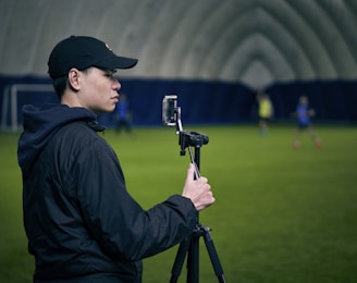 A person is standing indoors on a green field with a camera mounted on a tripod. The background appears to be a large, domed sports facility. The person is wearing a black cap and jacket and is focused on filming or taking a picture.