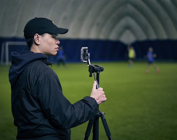 A person is standing indoors on a green field with a camera mounted on a tripod. The background appears to be a large, domed sports facility. The person is wearing a black cap and jacket and is focused on filming or taking a picture.