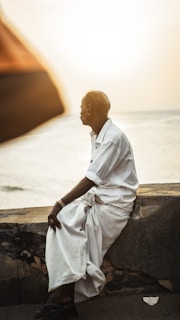 An elderly man in traditional attire sits on a stone structure, gazing towards the sea. The sun is setting in the background, casting a warm glow across the scene. The man's posture is relaxed, and his eyes are fixed on the distant horizon.