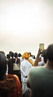 A group of people gathered outdoors, some facing away from the viewer. One individual is wearing a white outfit with an orange turban, holding a booklet. Another person is capturing the scene with a smartphone.