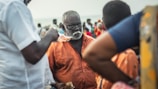 An older man with a white beard and glasses is wearing an orange shirt, surrounded by people. He is holding some currency, while another person is giving him money. The setting appears to be outdoors with a crowd in the background, suggesting a busy or market-like environment.