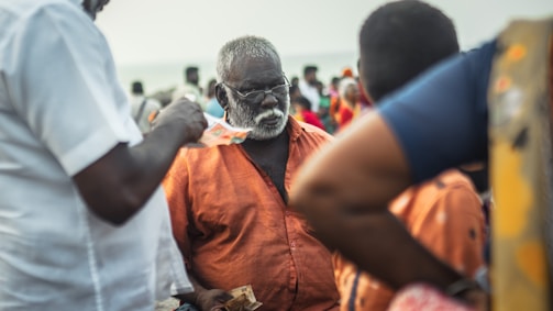 An older man with a white beard and glasses is wearing an orange shirt, surrounded by people. He is holding some currency, while another person is giving him money. The setting appears to be outdoors with a crowd in the background, suggesting a busy or market-like environment.