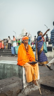 An elderly person dressed in a bright orange robe is seated on a concrete block in a bustling street. They hold a wooden staff and a bowl, with a serene expression. In the background, various people are walking, including a woman in a blue and gold saree. The scene is lively with social interaction and movement.