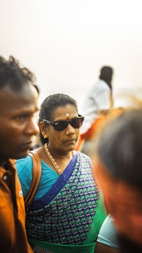 A woman wearing sunglasses is dressed in a vibrant blue and green traditional sari. She has a small white mark on her forehead and is accessorized with earrings and a necklace. She is surrounded by other people, one of whom is partially visible and wearing a brown shirt.