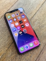 A smartphone lies on a wooden surface, displaying the home screen filled with various app icons. The screen appears cracked, especially noticeable in the bottom right corner.