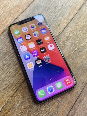 A smartphone lies on a wooden surface, displaying the home screen filled with various app icons. The screen appears cracked, especially noticeable in the bottom right corner.