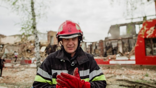 A firefighter wearing a red helmet and protective gear stands in front of a scene with rubble and damaged buildings. The surrounding area is in disarray, with trees and debris scattered around.