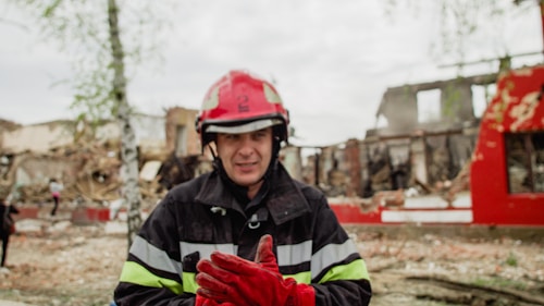 A firefighter wearing a red helmet and protective gear stands in front of a scene with rubble and damaged buildings. The surrounding area is in disarray, with trees and debris scattered around.