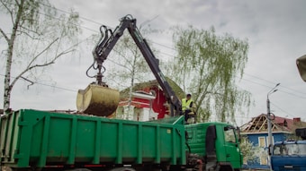 A construction vehicle features a large mechanical arm with a claw, lifting debris or soil into a green dump truck. The scene includes a worker wearing a high-visibility vest sitting in the vehicle. The surrounding environment includes trees with sparse leaves and partially visible buildings. The sky appears overcast.