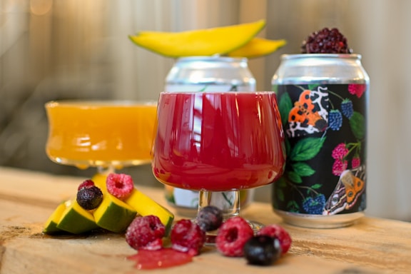 A close-up view of two colorful beverages served in decorative glasses, one red and one orange, alongside creatively designed cans. Fresh fruits, including raspberries, blackberries, and mango slices, are artfully placed around the drinks, with one glass having a slice of mango on its rim.