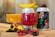 A close-up view of two colorful beverages served in decorative glasses, one red and one orange, alongside creatively designed cans. Fresh fruits, including raspberries, blackberries, and mango slices, are artfully placed around the drinks, with one glass having a slice of mango on its rim.