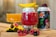 A close-up view of two colorful beverages served in decorative glasses, one red and one orange, alongside creatively designed cans. Fresh fruits, including raspberries, blackberries, and mango slices, are artfully placed around the drinks, with one glass having a slice of mango on its rim.