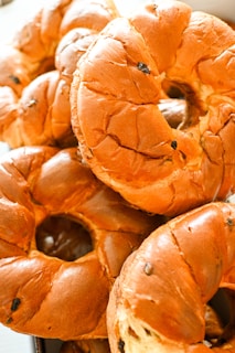 A close-up view of several freshly baked bagels, showcasing their golden-brown crusts and circular shape. The bagels have a glossy texture, with small spots indicating the presence of raisins or another similar ingredient.