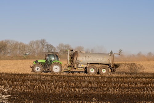 A green tractor is pulling a large tank trailer across a barren field. The trailer appears to be spreading fertilizer or compost onto the soil. In the background, there are leafless trees under a clear blue sky, indicative of late autumn or early spring.
