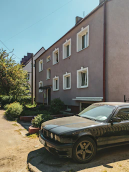 A large, beige residential building with multiple windows and a flat roof stands beside a small garden area. A black sedan is parked in the foreground, on a driveway next to the house. Bright sunlight casts shadows from the trees and building, and the sky is clear and blue.