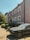 A large, beige residential building with multiple windows and a flat roof stands beside a small garden area. A black sedan is parked in the foreground, on a driveway next to the house. Bright sunlight casts shadows from the trees and building, and the sky is clear and blue.