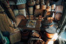 A wire basket hanging from the ceiling contains a wooden mousetrap placed centrally. Surrounding the basket are various household items including brooms, woven baskets, and wooden containers, creating a rustic and cluttered ambience.