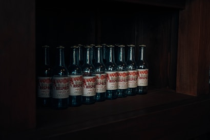 Several bottles with labels featuring red and white design are neatly arranged on a dark wooden shelf. The lighting is dim, creating a moody atmosphere.