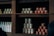 A wooden shelf is stocked with various canned and bottled goods. There are different brands and sizes of cans, some with red labels, others with beige and green labels. Bottled items wrapped in white paper are neatly lined up.
