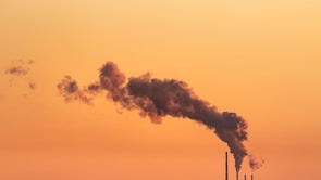 Thick dark smoke billows from industrial chimneys against a vibrant orange sky, likely during sunset or sunrise. The contrast between the industrial elements and the natural backdrop highlights themes of pollution and environmental impact.