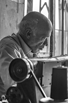 An elderly man focuses intently while working with a sewing machine in a room with soft natural light filtering through a window. His expression is one of concentration, and the setting has an industrial feel with visible gears and components.