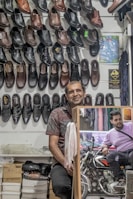 A man stands in a shoe store with an array of black and brown leather shoes displayed on the wall behind him. There are boxes of shoes on the shelf below. A mirror reflects another man sitting on a motorcycle outside the store.