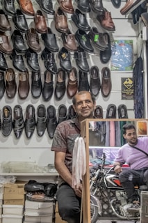 A man stands in a shoe store with an array of black and brown leather shoes displayed on the wall behind him. There are boxes of shoes on the shelf below. A mirror reflects another man sitting on a motorcycle outside the store.