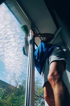 A person wearing a blue shirt and light-colored shorts is cleaning a large window. The sky outside is blue with scattered clouds and there are trees and parts of a building visible.