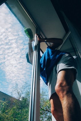 A person wearing a blue shirt and light-colored shorts is cleaning a large window. The sky outside is blue with scattered clouds and there are trees and parts of a building visible.
