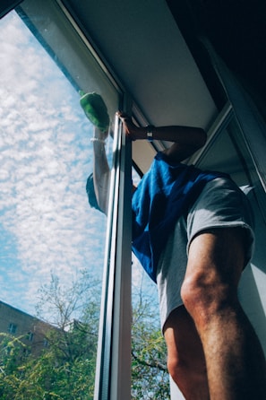 A person wearing a blue shirt and light-colored shorts is cleaning a large window. The sky outside is blue with scattered clouds and there are trees and parts of a building visible.