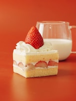 A square piece of strawberry shortcake featuring layers of vanilla sponge cake, cream, and sliced strawberries, topped with whipped cream and a whole strawberry. In the background, a clear glass filled with milk is placed on a warm orange surface.