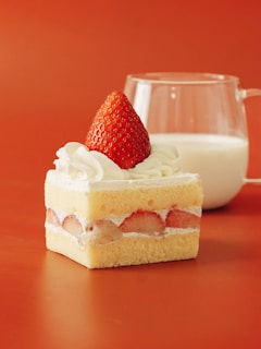 A square piece of strawberry shortcake featuring layers of vanilla sponge cake, cream, and sliced strawberries, topped with whipped cream and a whole strawberry. In the background, a clear glass filled with milk is placed on a warm orange surface.