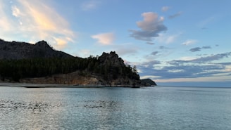 A serene coastal landscape with rocky cliffs in the foreground. Pine trees cover the cliffs, extending along the coastline. The sky is a blend of soft pastel colors with scattered clouds, reflecting on the calm waters below.