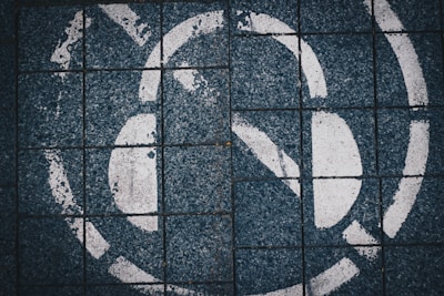A close-up view of a mosaic tiled surface featuring a large, partially visible white graffiti or painted symbol. The tiles are square and dark blue in color with prominent black lines in between, showing a worn texture with chipped areas.