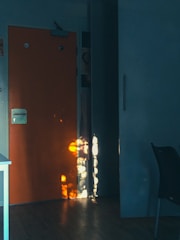 An interior scene featuring a wooden floor and a brightly lit wooden door with a metal handle. The door is contrasted against a dark wall, and rays of warm sunlight create patterns of reflected light on the surface, adding depth and interest to the composition. A small portion of a chair and a table edge are visible, suggesting this is a home or office space.