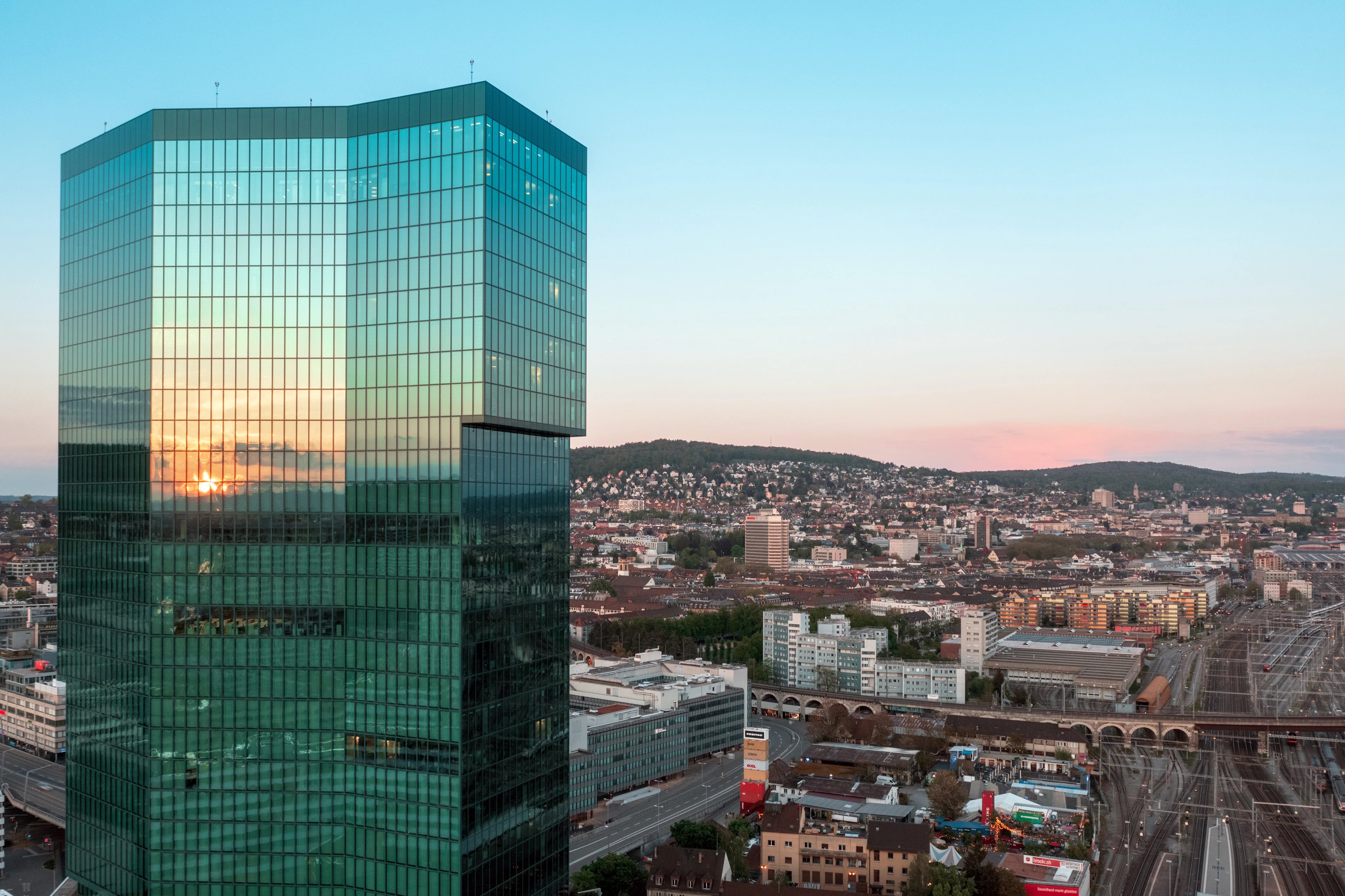 The green-glass Prime Tower rising over Zurich-West at sunset, the setting sun mirrored in its facade and the old railway viaduct, rail yards and low workshop roofs of the former factory quarter spread out below