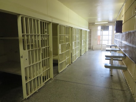 A corridor lined with prison cells, each secured with metal bars. The scene is empty and sterile with a row of metal tables attached to the wall.