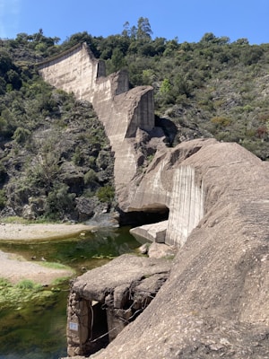 A broken dam structure made of concrete extends across a small valley, surrounded by lush green vegetation and trees. The remnants of the dam are partially collapsed, revealing jagged angles and exposed rebar. A shallow river runs beneath the structure, with clear water and patches of green algae.
