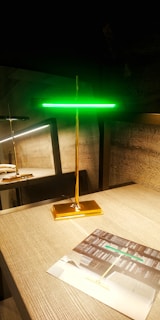 A modern, sleek lamp with a golden base and stem, topped by a glowing green horizontal light strip. It stands on a wooden tabletop next to a brochure displaying similar lighting designs. There is a reflection of the lamp on a glass surface behind it, enhancing the room's contemporary and stylish decor.