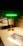 A modern, sleek lamp with a golden base and stem, topped by a glowing green horizontal light strip. It stands on a wooden tabletop next to a brochure displaying similar lighting designs. There is a reflection of the lamp on a glass surface behind it, enhancing the room's contemporary and stylish decor.